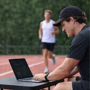 Coach working on a laptop next to a running track while an athlete trains in the background.